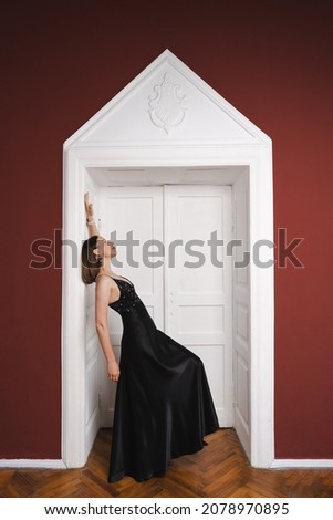 Beautiful woman model dressed in an elegant long black dress in a fashion pose in a modern interior with white door on burgundy wall