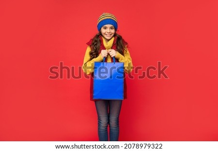 cheerful kid in winter hat hold shopping bags on red background, christmas shopping