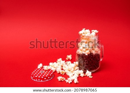 Full glass jar with handle, open. In the jar, coffee beans and popcorn fall out around. Red lid, white dots. Shadow. Red background. Text space. Celebration, drinks, food, concept. Minimal style.