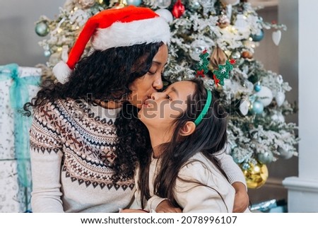 Black curly-haired mother in Santa hat kisses on cheek brunette daughter in holiday headband sitting against Christmas tree