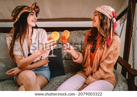 two carefree young women on vacation sitting on the rocking chair talking together and drinking fruit cocktails