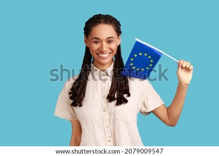 European Union flag. Adorable satisfied smiling woman with black dreadlocks holding Europe flag, looking at camera, wearing white shirt. Indoor studio shot isolated on blue background.