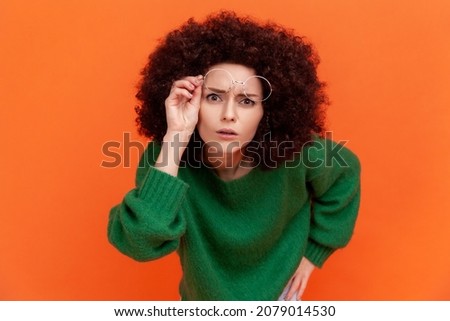 Curious woman with Afro hairstyle wearing green casual style sweater raising her optical glasses, wants to see something better, staring attentively. Indoor studio shot isolated on orange background. Royalty-Free Stock Photo #2079014530