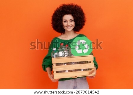 Satisfied woman with Afro hairstyle wearing green casual style sweater with box of empty plastic bottle and green sign, recycle, ecology concept. Indoor studio shot isolated on orange background.