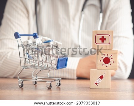 Vaccine bottles on a mini shopping trolley and health icons on wooden cubes on a wooden table with a doctor's sitting in the background. Vaccination and immunization. Medical and health care concept