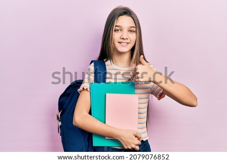 Young brunette girl holding student backpack and books smiling happy and positive, thumb up doing excellent and approval sign 