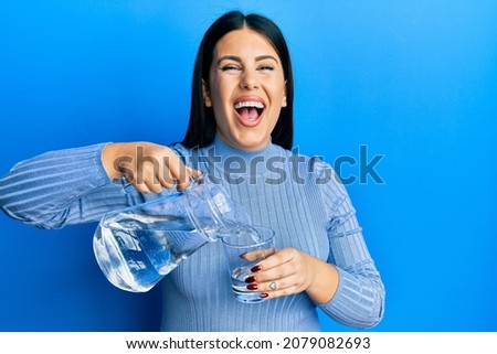 Beautiful brunette woman pouring water in glass smiling and laughing hard out loud because funny crazy joke. 
