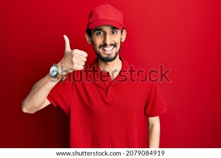 Young hispanic man wearing delivery uniform and cap smiling happy and positive, thumb up doing excellent and approval sign 