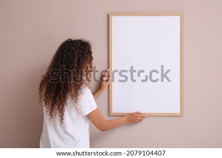 African American woman hanging empty frame on pale rose wall, back view. Mockup for design