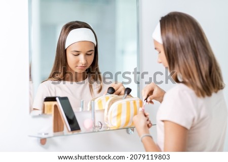 beauty, make up and cosmetics concept - teenage girl applying lip gloss and looking to mirror at home bathroom