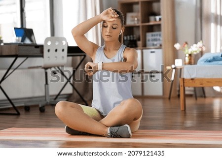sport, fitness and healthy lifestyle concept - tired teenage girl with smart watch and earphones sitting on yoga mat at home