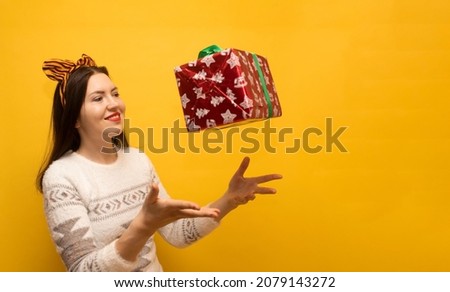 Beautiful smiling brunette in warm white sweater with deer and funny tiger ears on her head catches red Christmas gift on yellow background.Happy girl with new year flying present isolated.Copy space