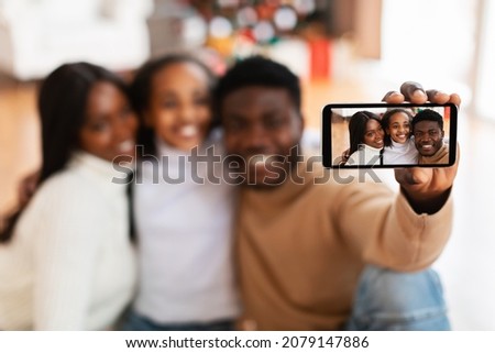 Capturing Moment. Happy loving black family of three taking selfie, posing at home on Xmas Eve, hugging and smiling at smartphone, selective focus on device gadget screen in hand, blurred background