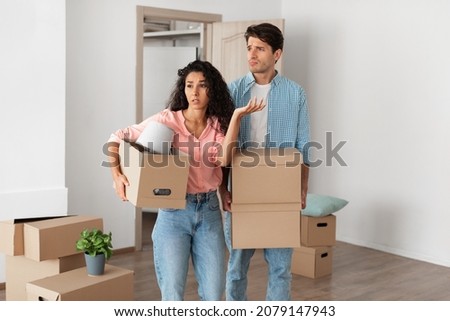 Portrait of disappointed young couple of newlyweds holding cardboard carton boxes walking in new flat, looking frustrated, young family moving into bad flat, sad people confused by interior design Royalty-Free Stock Photo #2079147943