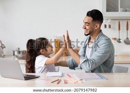 Handsome middle eastern man dad and adorable little girl child sitting at table, watching educational lesson on laptop, taking notes, celebrating success, giving each other high five, kitchen interior