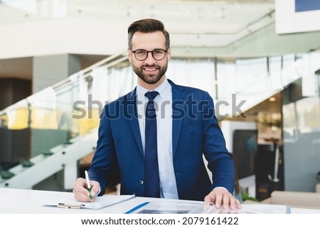 Successful caucasian smiling man shop assistant receptionist in formal attire writing looking at camera while standing at reception desk in hotel car dealer shop Royalty-Free Stock Photo #2079161422