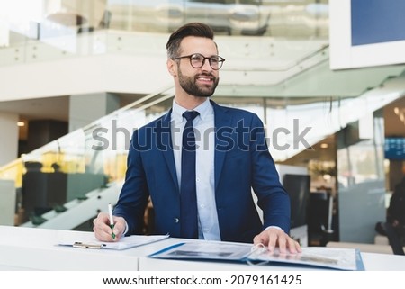 Successful caucasian smiling man shop assistant receptionist in formal attire writing while standing at reception desk in hotel car dealer shop Royalty-Free Stock Photo #2079161425