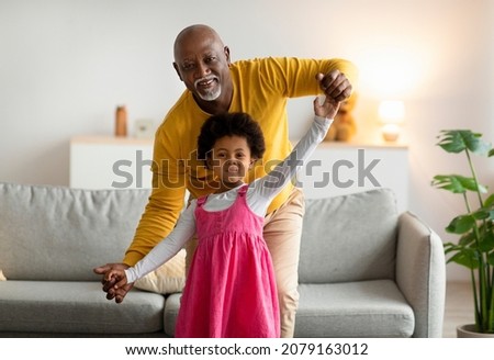 Smiling african american little girl and old man dancing and have fun in living room interior. Joy together, grandfather and granddaughter in free time at home. Grandpa visit, family, relationship Royalty-Free Stock Photo #2079163012