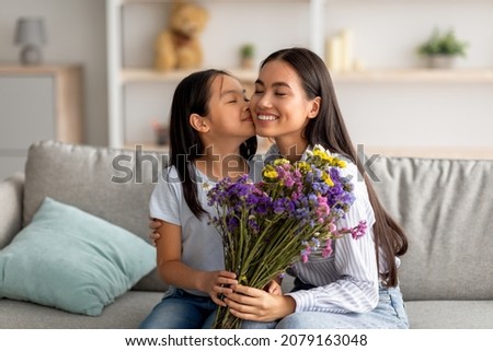 I love you, mommy. Pretty asian girl kissing her mum in cheek and giving bunch of flowers, greeting mom with holiday, Women's or Mother's day or birthday, sitting on sofa