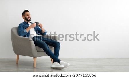 Full length of young Arab man drinking hot coffee in armchair against white studio wall, banner with free space. Peaceful middle Eastern guy having relaxing day, chilling on lazy morning Royalty-Free Stock Photo #2079163066