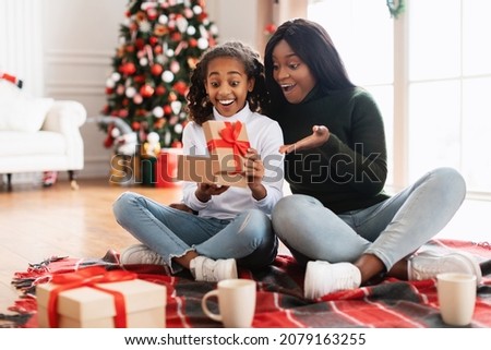 Winter Holiday Morning Concept. Happy black woman giving present to excited kid daughter, girl opening unwrapping Christmas gift sitting on floor blanket with decorated festive tree in living room