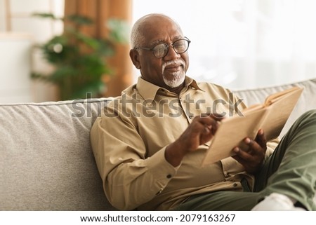 Senior African American Man Reading Book Sitting On Couch At Home, Wearing Eyeglasses. Retired Male Enjoying Reading New Novel Or Business Literature On Weekend. Retirement Leisure Concept Royalty-Free Stock Photo #2079163267