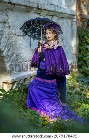A woman in a 19th century costume stands with a black umbrella near the ruins of an old castle