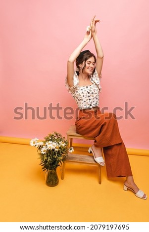 Full-length, elegant, fair-skinned young girl sitting on chair with her arms raised above head. Indoors, photo of pretty brown-haired woman in white blouse, red pants and shoes. Royalty-Free Stock Photo #2079197695