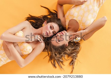 Young beautiful european women at photo shoot in studio, posing tenderly for camera. Fair-skinned girls with wavy hair in swimsuits on yellow background.