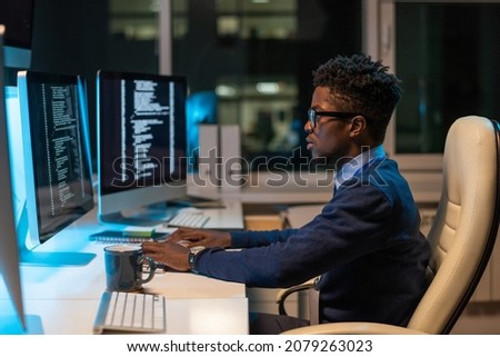 Young it-engineer of African ethnicity working with coded data while sitting in armchair in front of computer in office Royalty-Free Stock Photo #2079263023
