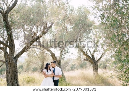 Groom hugs bride while standing between trees in a green park