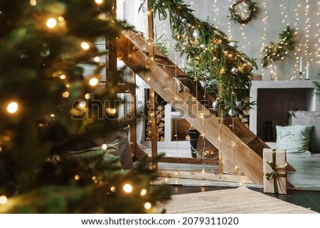 Home New Year's interior. A live Christmas tree and a wooden staircase in the kitchen. Cozy atmosphere of a home holiday, loft design of the room, daylight, green tree decorated with lights