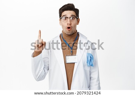 Amazed doctor in glasses and robe pointing up, showing big promo, hospital news, making announcement, standing over white background Royalty-Free Stock Photo #2079321424