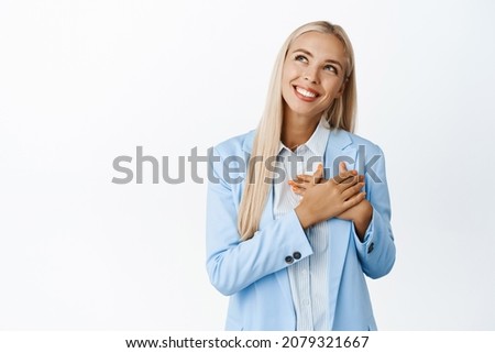 Dreamy businesswoman, holding hands on heart, smiling and gazing aside, imaging smth, picturing romantic moment, standing over white background
