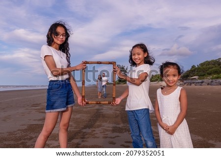 family, leisure and people concept-asian happy female siblings holding picture of frame of parents on the beach