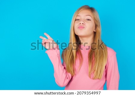 caucasian little kid girl wearing long sleeve shirt over blue background makes peace gesture keeps lips folded shows v sign. Body language concept