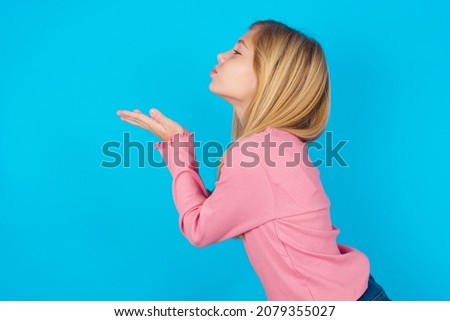 Profile side view view portrait of attractive caucasian little kid girl wearing long sleeve shirt over blue background sending air kiss