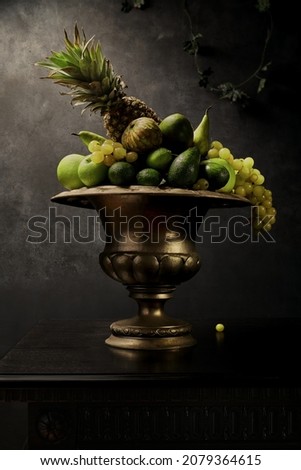 Still Life with many different green fruits in big silver vase. Dark wall background, antique wooden table, pineapple, wine grapes, green apples, limes. Great for designers and interior decoration. 