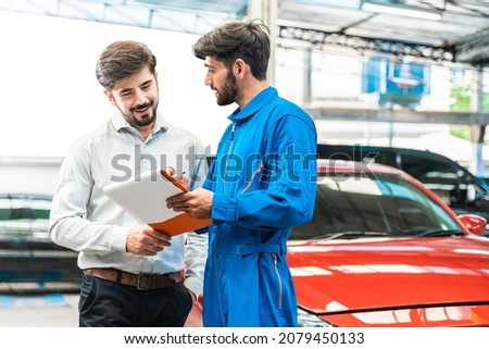 Mechanic show the car checking list to customer before giving him a car key with the blur lifted car in the garage. Focus on customer. Auto car repair service center. Professional service. Royalty-Free Stock Photo #2079450133