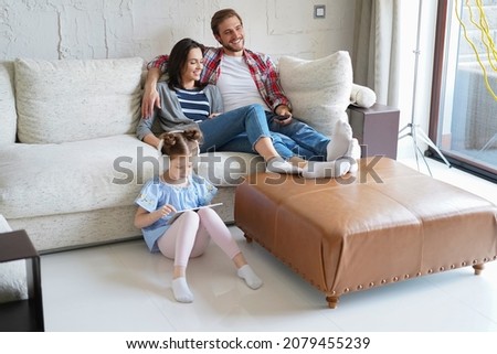 Smiling happy family sit relax on couch in living room watch little daughter drawing in digital tablet. Happy weekends at home