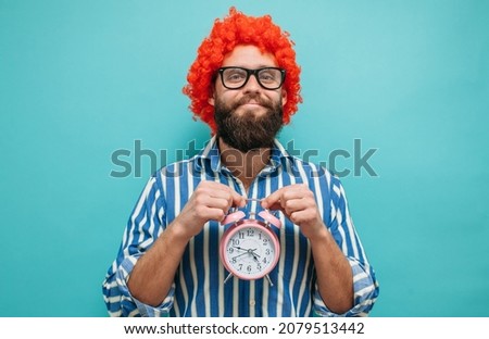 Young charismatic hipster with red hair holding alarm clock in hands on blue isolated background. Crazy emotions. It's time