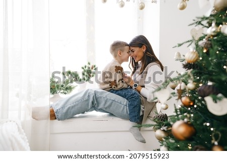 Happy mother with son sit on windowsill near Christmas tree
