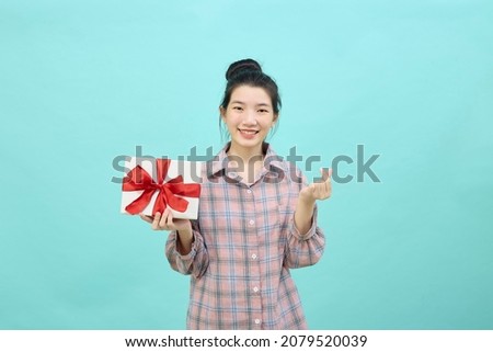 Charming young woman Asian with dark long hair feeling happy smiling to the camera and makes Korean love sign shapes heart, with fingers and holding gift box isolated over blue background.