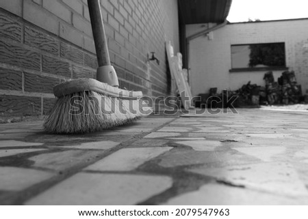A black and white portrait of the brush of a broom standing on a pavement next to a house.