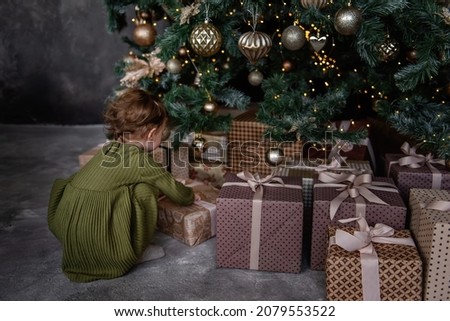 Little girl in green warm dress opens boxes of gifts in craft paper under a Christmas tree. Elegant decor with golden balls, toys. Retro style in a gray loft interior. Home decoration for the holiday