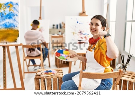 Young artist woman painting on canvas at art studio smiling doing phone gesture with hand and fingers like talking on the telephone. communicating concepts. 