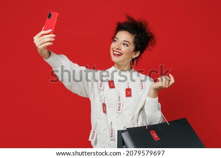 Young smiling happy woman 20s wear white knitted sweater with tags sale in store showroom doing selfie shot on mobile cell phone post photo on social network isolated on plain red background studio