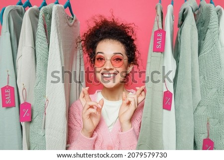 Young costumer woman in sweater glasses stand near clothes rack with tag sale in store showroom waiting for special moment, keep fingers crossed, making wish isolated on plain pink background studio