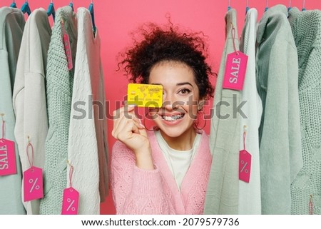 Young smiling fun female costumer woman 20s wear sweater stand near clothes rack with tag sale in store showroom hold cover eye with credit bank card isolated on plain pink background studio portrait