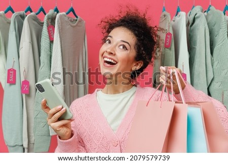 Young minded fun woman in sweater stand near clothes rack with tag sale in store showroom hold package bags with purchases after shopping mobile cell phone isolated on plain pink background studio.